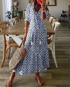 Robe longue bohème bleue et blanche à motifs, femme tenant un sac en osier, sandales marron, intérieur moderne avec table en bois et chaises.