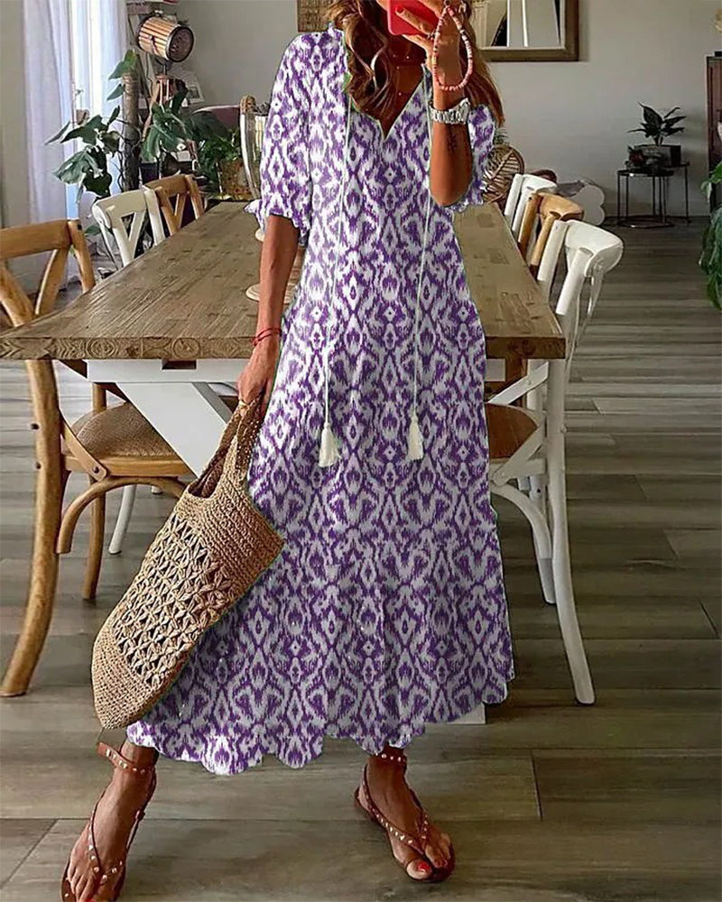 Robe longue bohème violette à motifs géométriques, femme tenant un sac en osier, sandales marron, intérieur moderne avec table en bois.