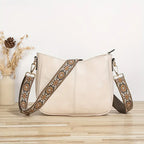 Sac à main en cuir beige avec bandoulière brodée florale, posé sur une table en bois clair, vase décoratif en arrière-plan. Accessoire mode élégant.