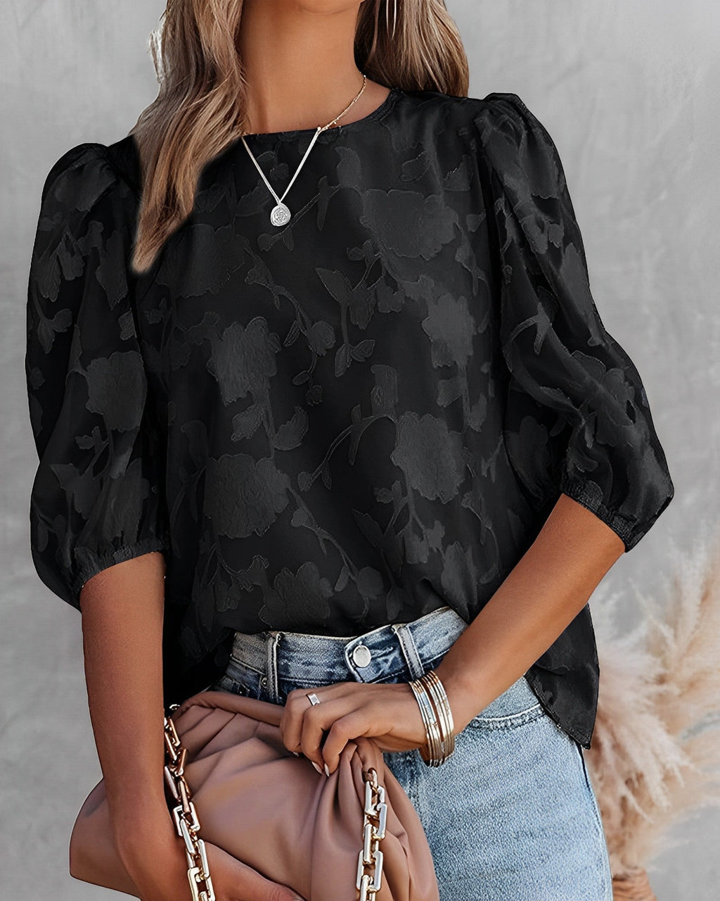 Femme portant un chemisier noir à motifs floraux, manches bouffantes, avec un jean bleu et un sac beige. Mode élégante et tendance.