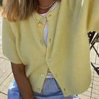 Femme portant un cardigan jaune en maille avec boutons, collier de perles et pendentif doré, assise sur une terrasse ensoleillée. Mode été chic.
