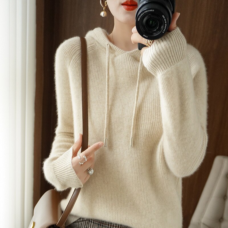 Femme portant un pull en laine beige avec capuche, tenant un appareil photo. Mode automne-hiver, style décontracté, accessoires élégants.