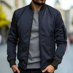Homme portant un blouson aviateur bleu marine, t-shirt gris, dans une rue urbaine floue. Mode masculine, style décontracté, tendance automne.