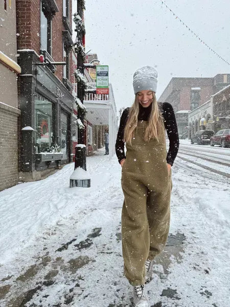 Jeune femme souriante en salopette marron et bonnet blanc marchant dans une rue enneigée, devant des boutiques, ambiance hivernale urbaine.