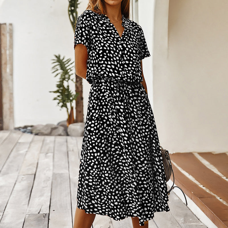 Robe noire à pois blancs, manches courtes, portée par une femme sur une terrasse en bois. Mode estivale élégante et décontractée.