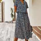 Robe midi à pois noirs et blancs, manches courtes, portée par une femme sur une terrasse en bois, avec plantes en arrière-plan.