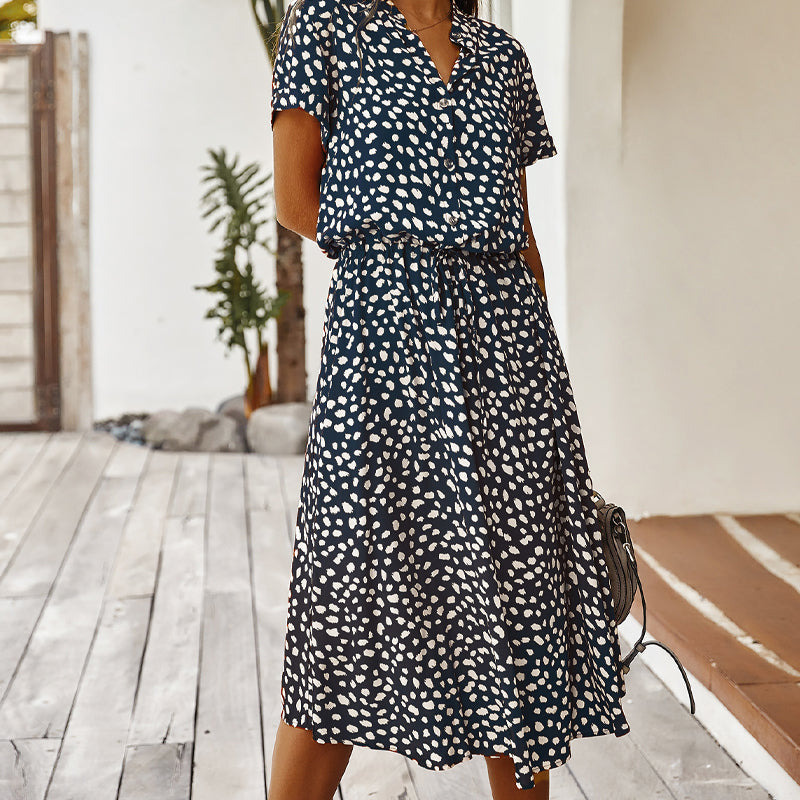 Robe midi à pois noirs et blancs, manches courtes, portée par une femme sur une terrasse en bois, avec plantes en arrière-plan.