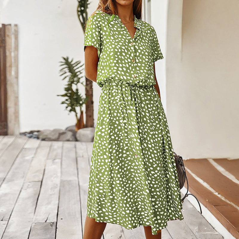 Robe verte à pois blancs, manches courtes, col en V, portée par une femme sur une terrasse en bois. Mode estivale, tenue décontractée élégante.