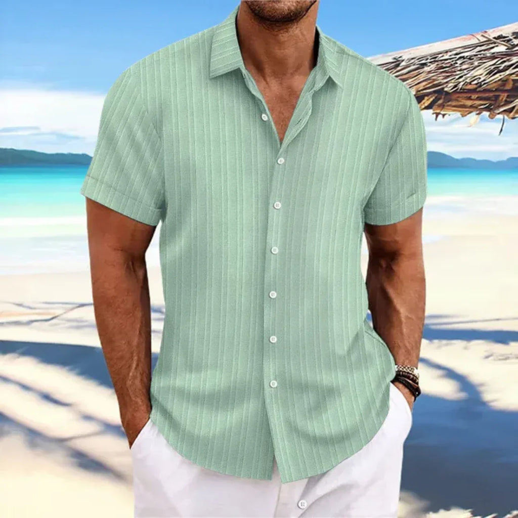 Homme en chemise verte à rayures sur plage tropicale, ciel bleu et mer en arrière-plan. Mode estivale, vacances, style décontracté.