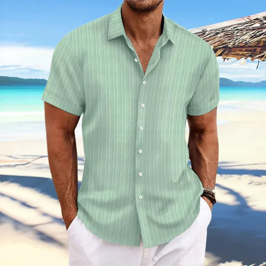 Homme en chemise verte à rayures sur plage tropicale, ciel bleu et mer en arrière-plan. Mode estivale, vacances, style décontracté.