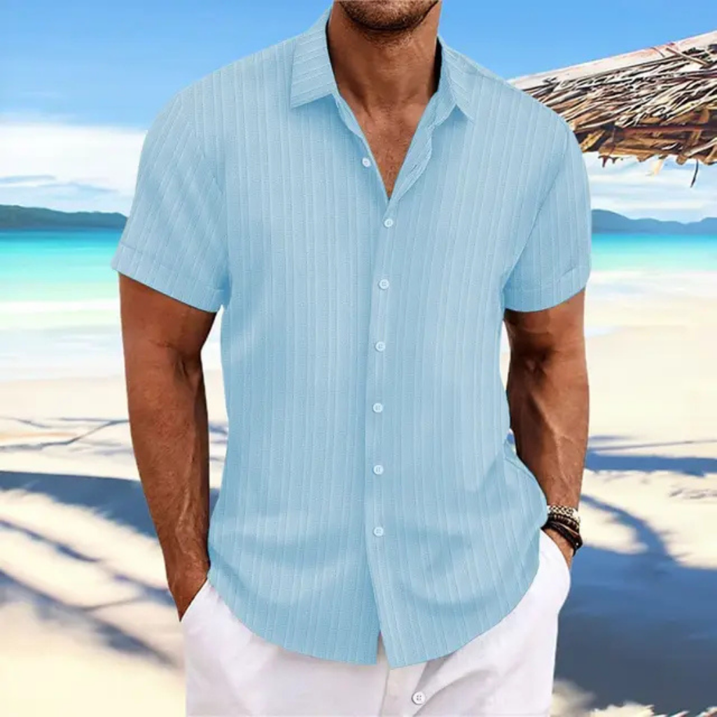 Homme en chemise bleue à rayures sur plage tropicale, mer turquoise en arrière-plan, ciel bleu, tenue estivale décontractée, vacances bord de mer.