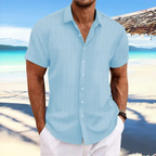 Homme en chemise bleue à rayures sur plage tropicale, mer turquoise en arrière-plan, ciel bleu, tenue estivale décontractée, vacances bord de mer.