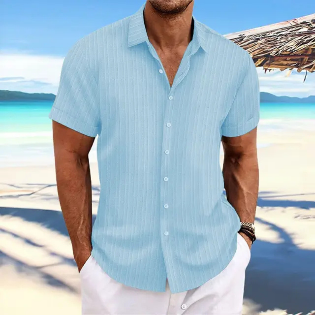 Homme en chemise bleue à rayures sur plage tropicale, mer turquoise en arrière-plan, ciel bleu, tenue estivale décontractée, vacances bord de mer.