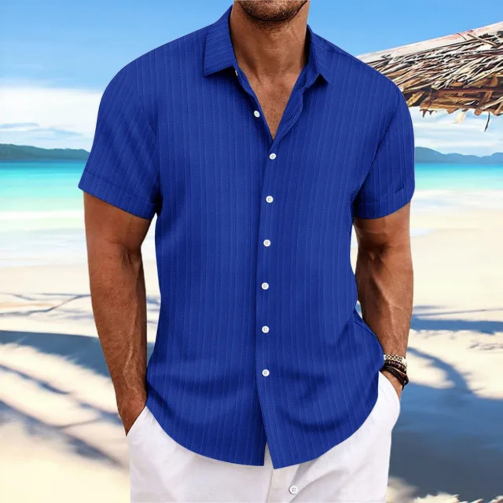 Homme en chemise bleue à manches courtes sur une plage tropicale, mer turquoise en arrière-plan, ciel bleu, ambiance estivale et décontractée.