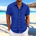Homme en chemise bleue à manches courtes sur une plage tropicale, mer turquoise en arrière-plan, ciel bleu, ambiance estivale et décontractée.