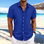 Homme en chemise bleue à manches courtes sur une plage tropicale, mer turquoise en arrière-plan, ciel bleu, ambiance estivale et décontractée.