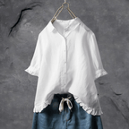 Chemisier blanc élégant avec manches courtes et volants, suspendu sur un cintre, sur fond gris texturé. Mode féminine, style décontracté.