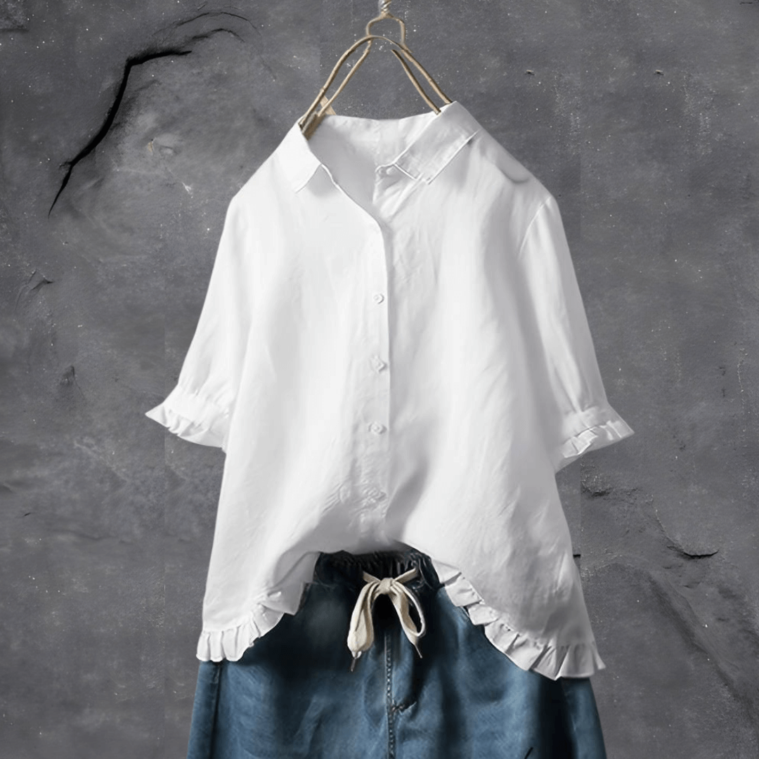Chemisier blanc élégant avec manches courtes et volants, suspendu sur un cintre, sur fond gris texturé. Mode féminine, style décontracté.