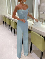 Femme en combinaison bleu ciel élégante avec plumes, sac à main blanc, debout dans une salle moderne avec chaises vertes et miroir.