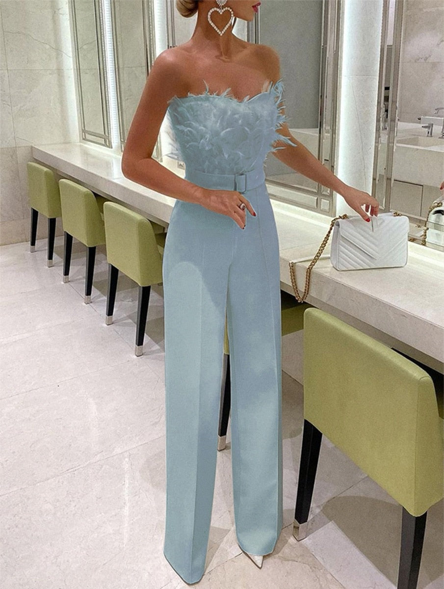 Femme en combinaison bleu ciel élégante avec plumes, sac à main blanc, debout dans une salle moderne avec chaises vertes et miroir.