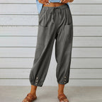 Pantalon ample gris pour femme avec poches avant, taille élastique et boutons décoratifs aux chevilles, style décontracté, fond bois clair.