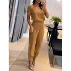 Femme portant une combinaison marron élégante, debout dans un salon moderne avec table noire et plante verte. Mode féminine chic et décontractée.