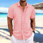 Homme en chemise rose à rayures sur plage tropicale, mer turquoise en arrière-plan, ciel bleu, ambiance estivale, mode décontractée.