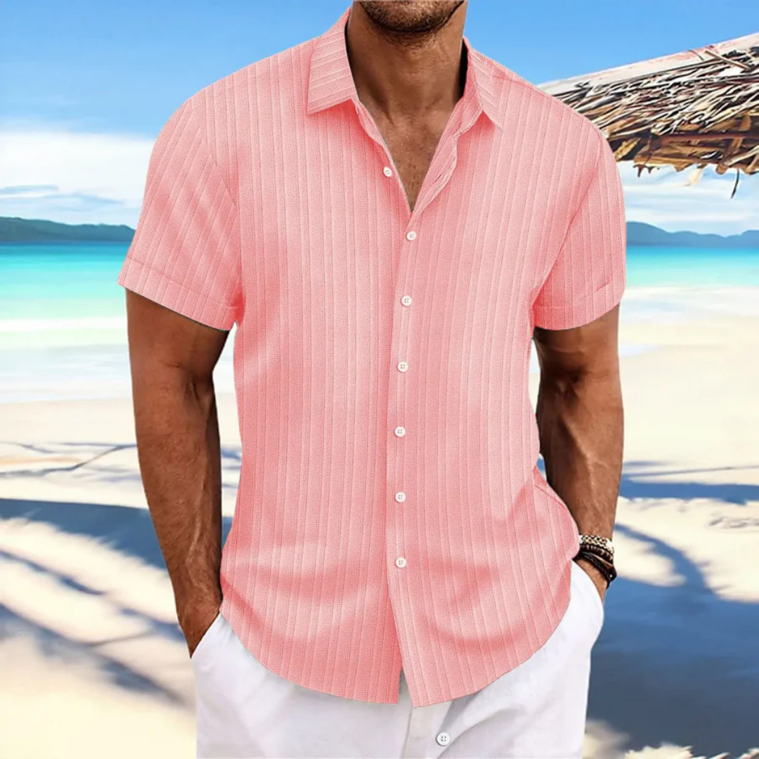 Homme en chemise rose à rayures sur plage tropicale, mer turquoise en arrière-plan, ciel bleu, ambiance estivale, mode décontractée.