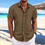 Homme en chemise marron à rayures, manches courtes, sur une plage tropicale avec mer turquoise et ciel bleu. Mode estivale, vacances, style décontracté.