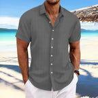 Homme en chemise grise à rayures, manches courtes, sur une plage tropicale avec mer turquoise et ciel bleu. Mode estivale, vacances plage.