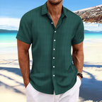 Homme en chemise verte à rayures, manches courtes, sur une plage tropicale avec mer turquoise et ciel bleu, style estival décontracté.