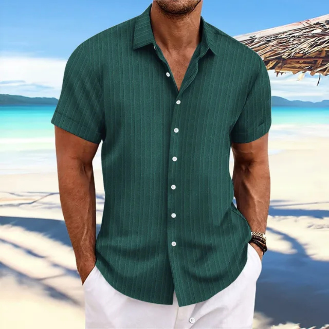 Homme en chemise verte à rayures, manches courtes, sur une plage tropicale avec mer turquoise et ciel bleu, style estival décontracté.