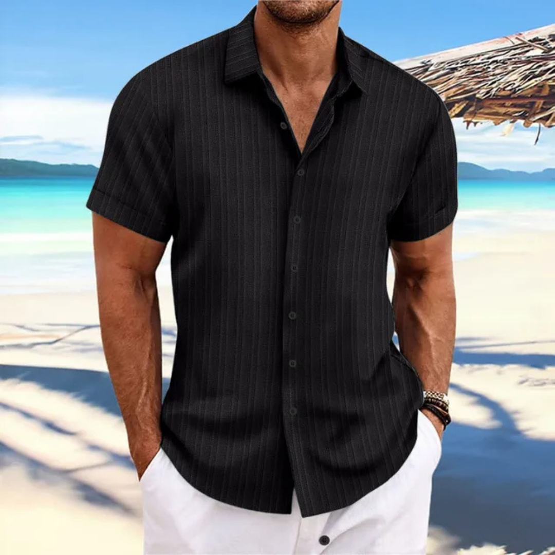 Homme en chemise noire à rayures, manches courtes, sur une plage tropicale avec mer turquoise et ciel bleu, style estival décontracté.