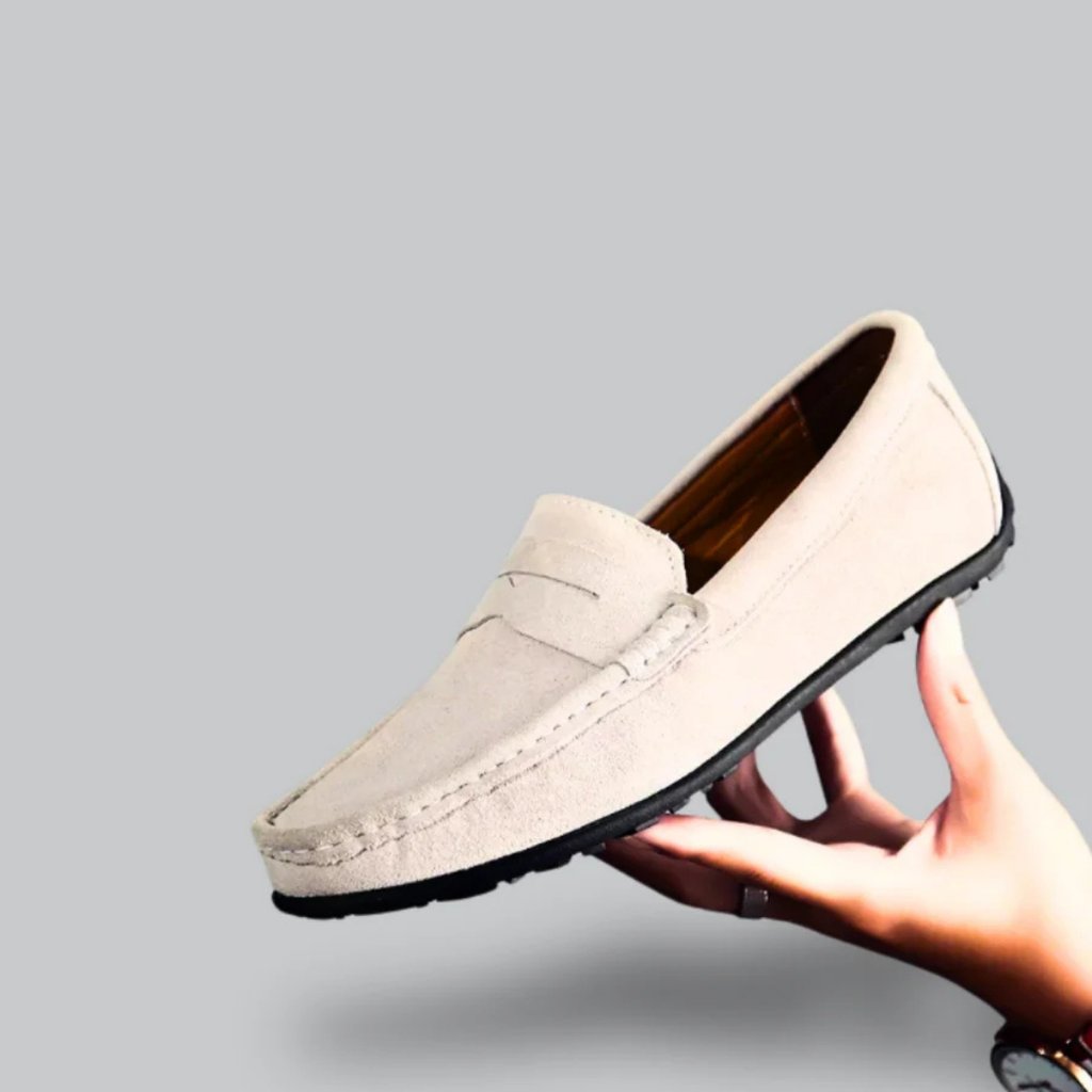 Mocassin beige en daim pour homme, semelle noire, tenu par une main. Chaussure élégante et confortable, idéale pour un style décontracté.