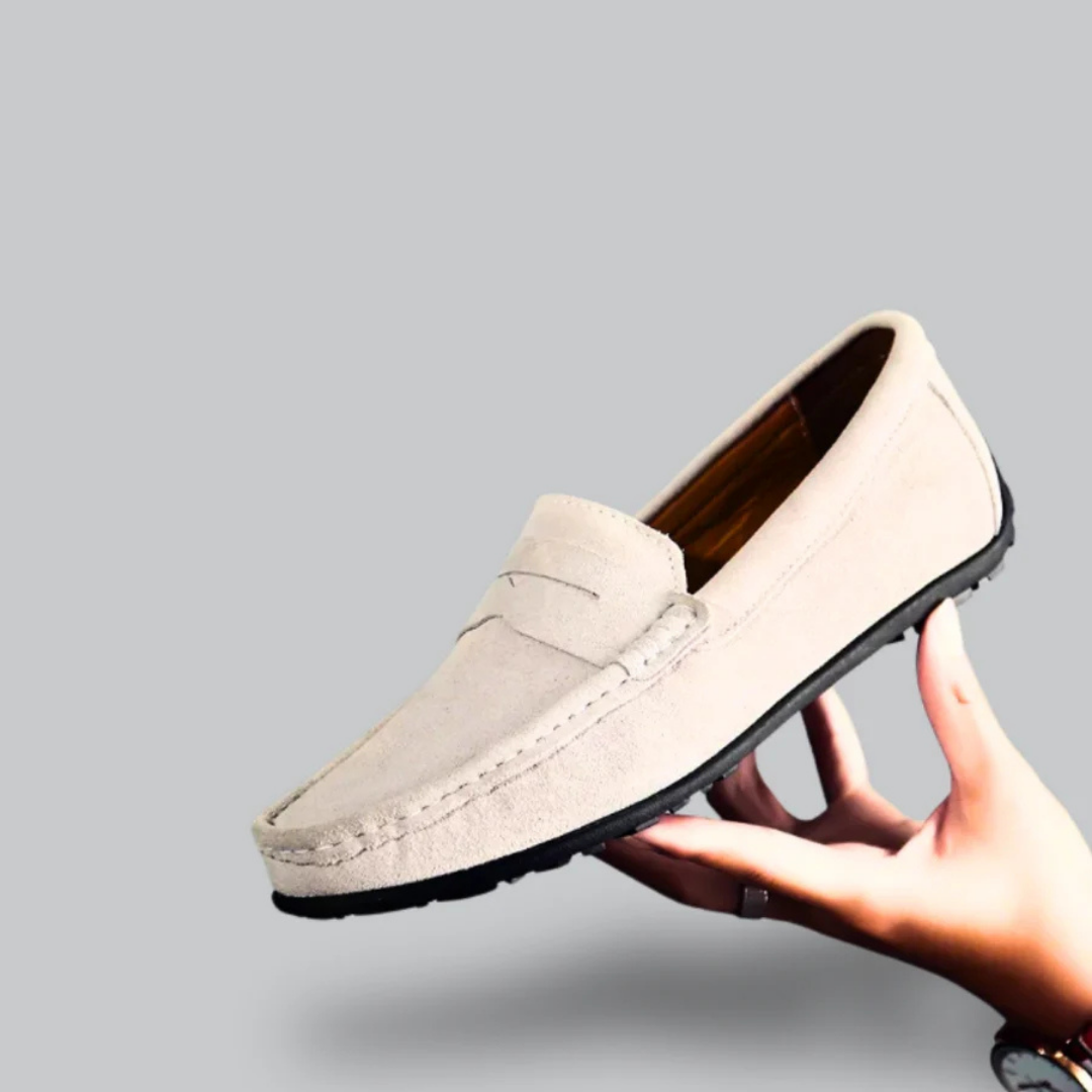 Mocassin beige en daim pour homme, semelle noire, tenu par une main. Chaussure élégante et confortable, idéale pour un style décontracté.
