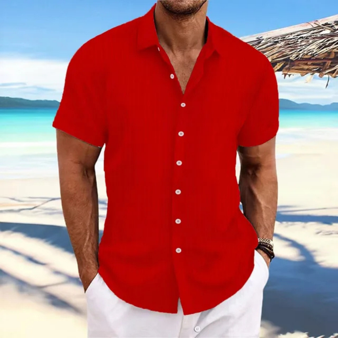 Homme en chemise rouge à manches courtes sur une plage tropicale, ciel bleu et mer en arrière-plan, style estival décontracté.