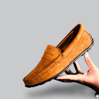 Mocassin en daim marron pour homme, semelle noire, tenu par une main sur fond gris. Chaussure élégante, style décontracté, mode masculine.