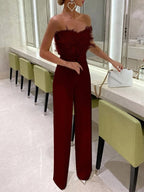 Femme en combinaison rouge élégante avec plumes, sac à main blanc, debout dans une salle moderne avec chaises vertes et miroir. Mode soirée chic.