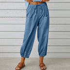 Pantalon bleu décontracté pour femme avec poches avant, boutons décoratifs aux chevilles, porté avec des sandales marron sur fond de mur en bois blanc.