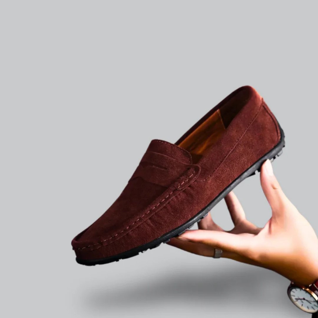 Mocassin en daim marron pour homme, semelle noire, tenu par une main. Chaussure élégante et confortable, idéale pour un style décontracté.
