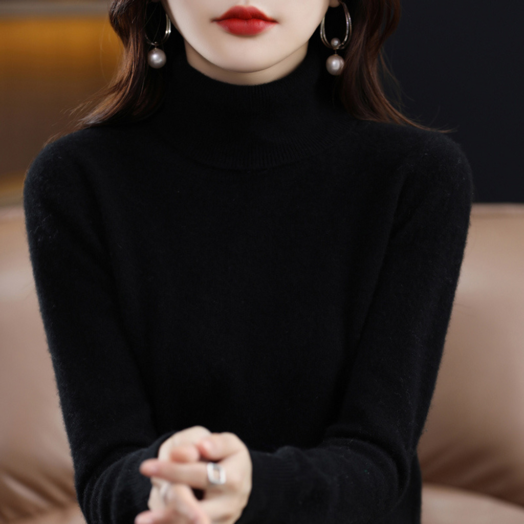 Femme en pull noir à col roulé, portant des boucles d'oreilles pendantes avec perles, assise sur un canapé beige. Mode élégante et moderne.