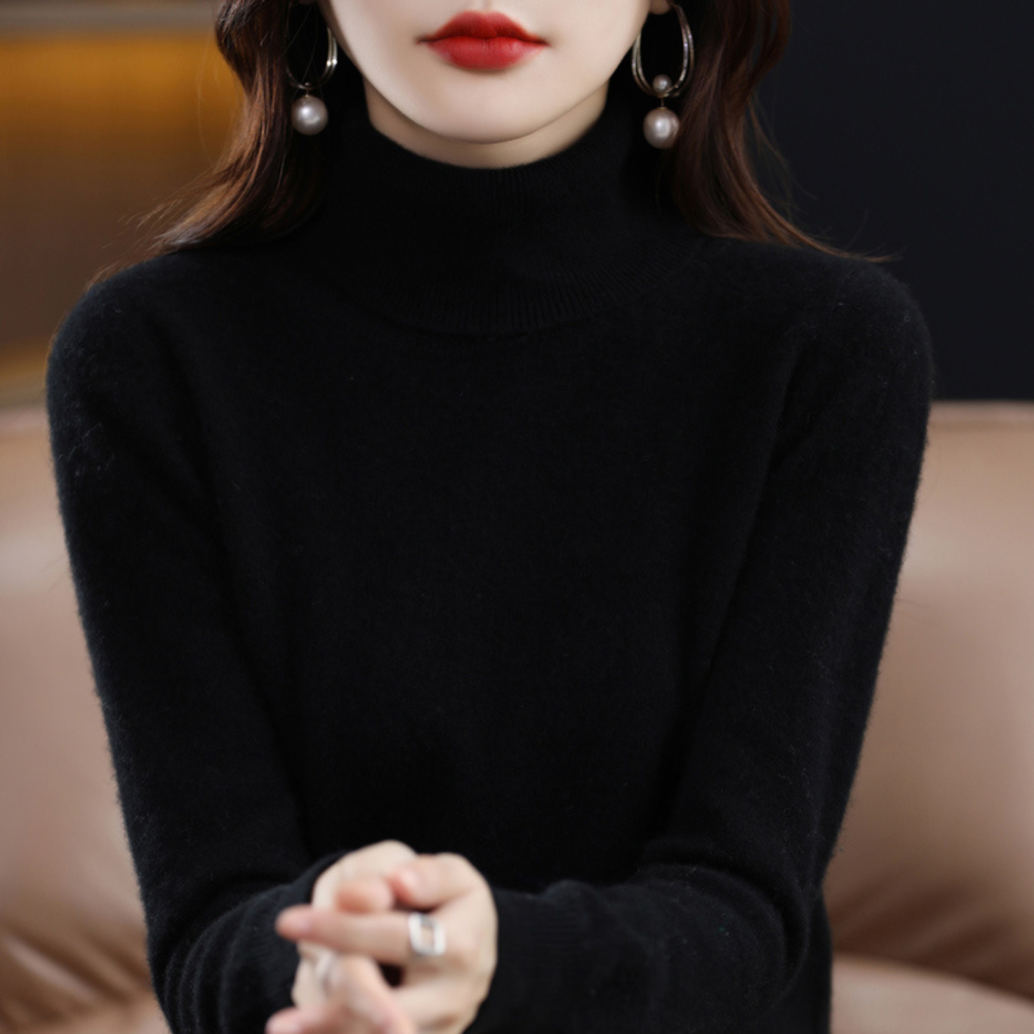 Femme en pull noir à col roulé, portant des boucles d'oreilles pendantes avec perles, assise sur un canapé beige. Mode élégante et moderne.