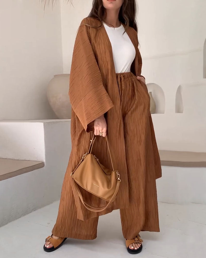 Femme portant un ensemble kimono marron plissé avec un sac à main assorti, sandales et t-shirt blanc, posant dans un intérieur minimaliste.