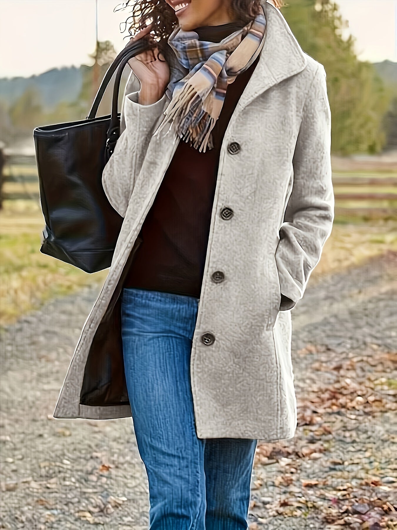 Femme souriante en manteau beige, écharpe à carreaux, sac noir, marchant sur un chemin rural. Mode automne, style décontracté, plein air.