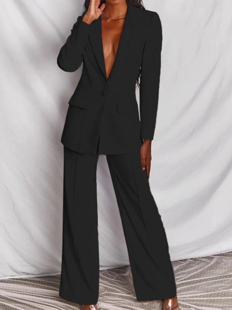 Femme portant un élégant costume noir, veste à col profond et pantalon large, posant devant un fond blanc. Mode chic et moderne.
