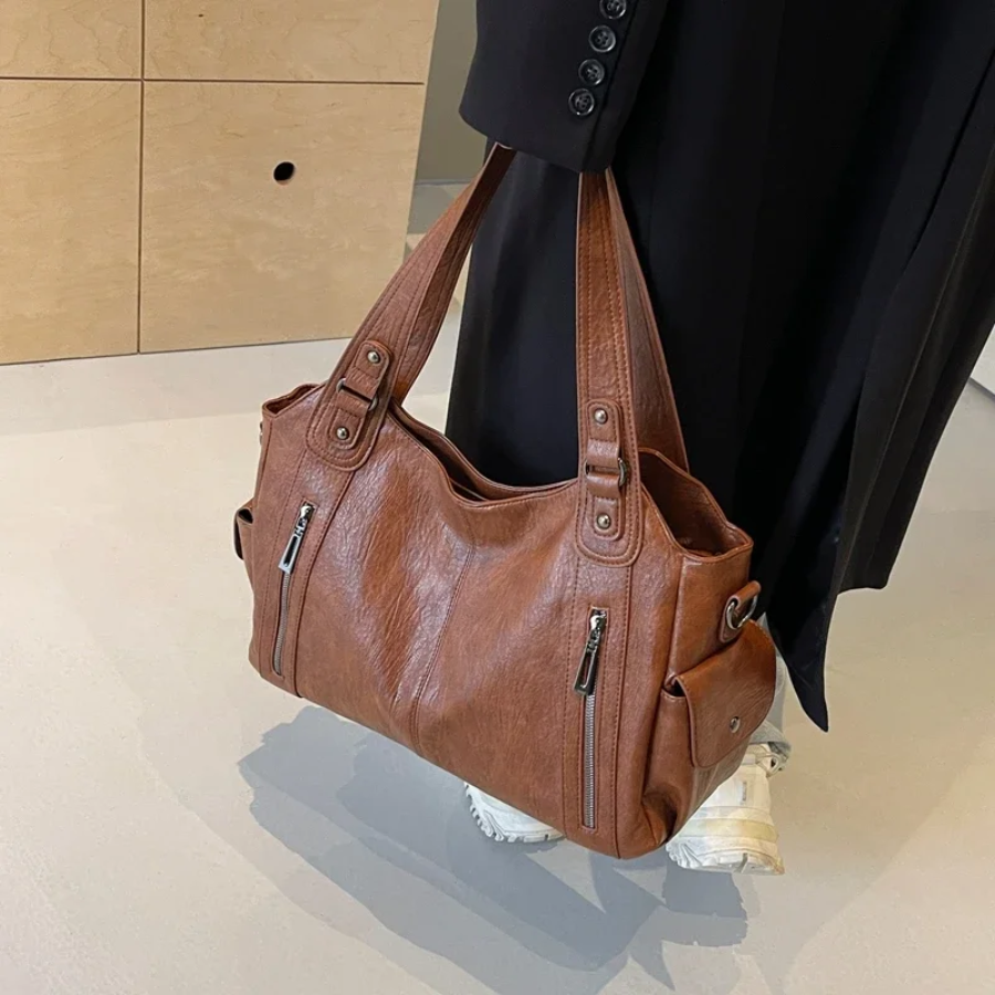 Sac à main en cuir marron avec poches zippées, porté par une personne en manteau noir et baskets blanches, sur fond de sol clair et mur en bois.
