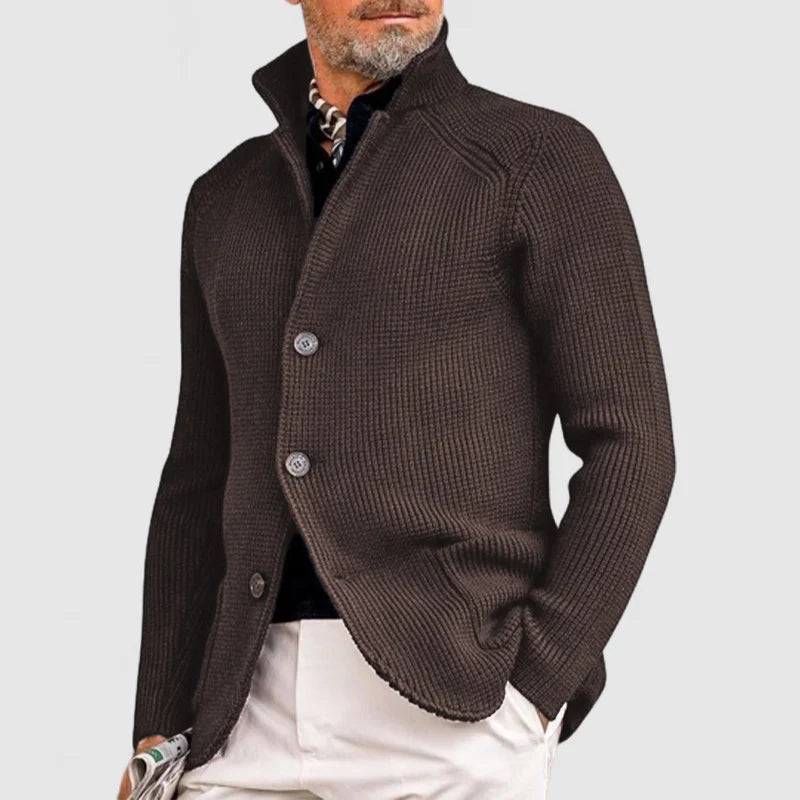 Homme portant un cardigan marron élégant, col relevé, boutons visibles, sur chemise noire, pantalon blanc. Mode masculine chic, style automne-hiver.