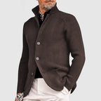 Homme portant un cardigan marron élégant, col relevé, boutons visibles, sur chemise noire, pantalon blanc. Mode masculine chic, style automne-hiver.