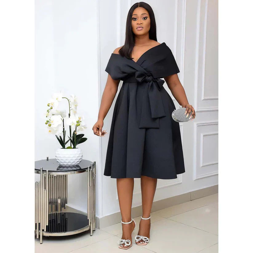 Femme élégante en robe noire chic, tenant une pochette argentée, debout près d'une table moderne avec orchidée blanche, intérieur sophistiqué.