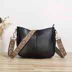 Sac à main noir en cuir avec bandoulière brodée florale marron, posé sur une table en bois clair. Accessoire mode élégant et tendance.